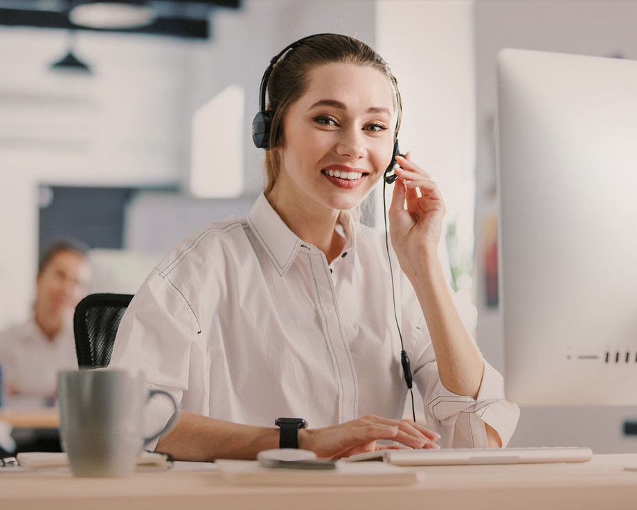 Eine Frau sitzt an einem Schreibtisch und spricht in ein Headset, während sie auf einem Computer arbeitet.